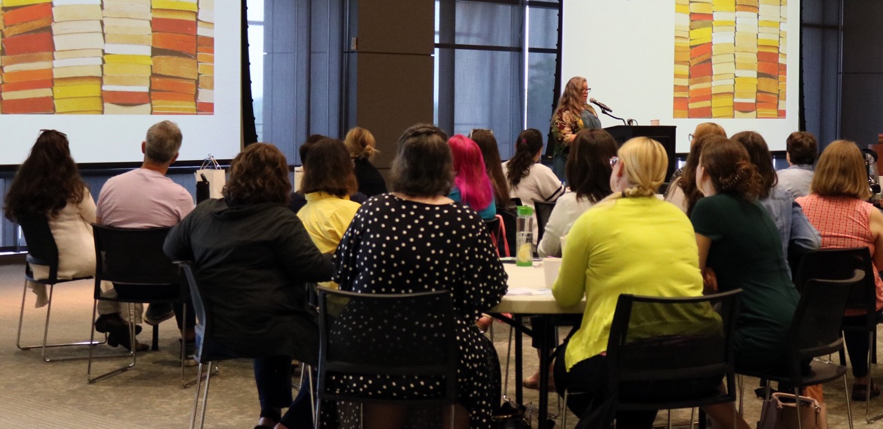 A room of librarians seated at tables. They are all facing Rebecca Tolley, who is giving a talk at a podium.